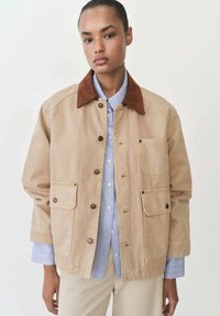 Beige canvas jacket with a brown suede collar, button front, two chest pockets, and a relaxed fit over a light blue shirt.