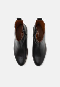 Černé kožené kotníkové boty s hladkým povrchem, špičatým tvarem, bočními zipy a hnědou oblastí podšívky.