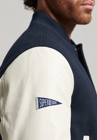 Marinblå varsityjacka med vita lädersleeves. Har ribbad krage och marinblå detaljer, med en broderad "Superdry"-logotyp på ärmen.