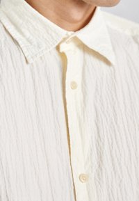 Valkoinen teksturoitu napitettava paita pehmeästä, ryppyisestä kankaasta. Ominaisuuksina terävä kaulus ja kevyet, läpikuultavat napit.