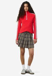Felpa rossa con zip e strisce bianche, abbinata a una gonna a pieghe a quadri grigia e verde, indossata con scarpe da ginnastica grigie e calzini bianchi.
