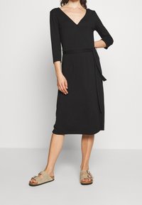 Svart omslagklänning med V-neck, trekvartslånga ärmar och ett knytbälte; knälång med en slät tygkvalitet. Bärs med beige sandaler.