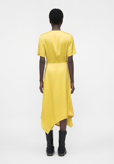 Άτομο με κοντά σγουρά μαλλιά που φοράει ένα κίτρινο φόρεμα με ασύμμετρη στρίφωμα και μαύρες μπότες μέχρι τη μέση της γάμπας, στέκεται μπροστά σε ένα απλό φωτεινό φόντο.