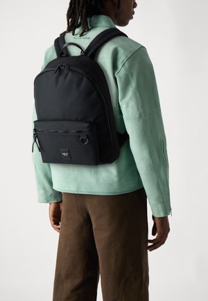 Sac à dos noir en matériau texturé, avec une poche zippée à l'avant et des bretelles réglables, porté sur une veste vert menthe.
