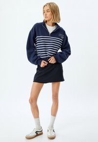 Sudadera de color azul marino con cremallera y rayas horizontales blancas, con puños y cuello acanalados. Combinada con una falda corta negra y zapatillas blancas.