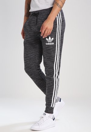 Person iført mørkegrå Adidas træningsbukser med hvide striber og logo, kombineret med hvide sneakers og en lysegrå skjorte.