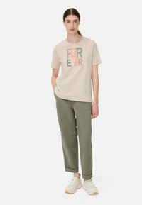 T-shirt beige clair avec le texte coloré "FOREVER", associé à un pantalon vert olive. Le modèle porte des baskets légères, les mains dans les poches, fond neutre.
