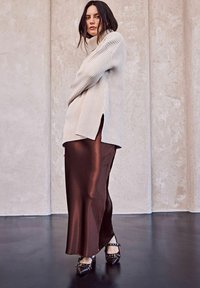 Maglione beige a coste oversize con collo alto e spacchi laterali, abbinato a una gonna in raso marrone lucido. Scarpe nere a punta con dettagli borchiati.