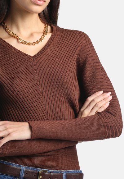 Femme portant un pull marron côtelé à col en V, un collier chaîne en or et un jean bleu avec une ceinture marron, bras croisés sur la poitrine.