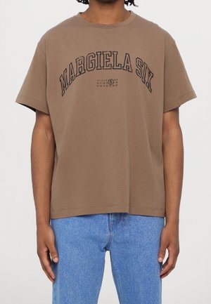 Osoba ubrana w brązowy t-shirt z krótkim rękawem z napisem "MARGIELA SIX" i logo z cyfrą 6 w kółku, w połączeniu z niebieskimi dżinsami.