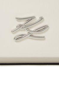 Biela kožená plocha s čisto strieborným kovovým logom, ktoré sa vyznačuje plynulými, preplétajúcimi sa líniami. Štruktúra vyzerá hladko a elegantne.