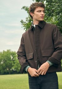 Chaqueta externa marrón con un collar de pana, dos bolsillos frontales y mangas texturizadas sobre una camisa verde clara, llevada por un hombre en un entorno natural.