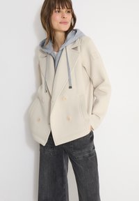 Femme portant une veste beige à double boutonnage sur un sweat à capuche gris, associée à un pantalon cargo noir, debout contre un mur blanc.