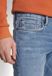 Světle modré džíny z denimu s opotřebovanými detaily, pětikapsovým designem a jemným prošíváním. Kombinace s oranžovým žebrovaným svetrem.