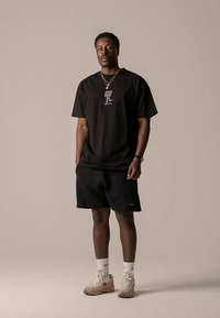 Čierne tričko s potlačou vpredu, doplnené o čierne šortky. Béžové tenisky s textúrovanými detailmi. Jednoduché pozadie.