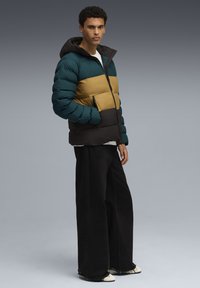 Jeune homme debout portant une veste rembourrée en blocs de couleurs avec des sections teal, moutarde et noire, un pantalon noir à jambes larges et des baskets blanches.