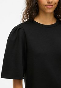 Črna majica s kratkimi rokavi in puffastimi rokavi, izdelana iz gladkega materiala. Okrogla izrez z našitimi detajli ob robu.