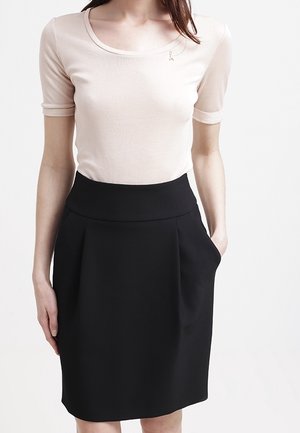 Femme portant un haut beige à manches courtes rentré dans une jupe noire taille haute mi-longue avec des poches, debout devant un fond uni.