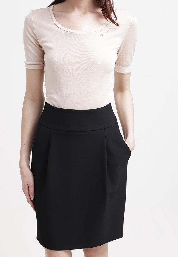 Femme portant un haut beige à manches courtes rentré dans une jupe noire taille haute mi-longue avec des poches, debout devant un fond uni.