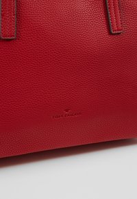 Červená kožená kabelka s texturou a dvěma uchy. Obsahuje embosované logo "TOM TAILOR". Hladký povrch, robustní design.