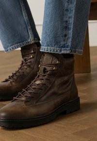 Bottines en cuir brun avec une texture lisse, dotées de lacets marron foncé, d'œillets en métal et d'une semelle en caoutchouc, portées avec un jean en denim clair.