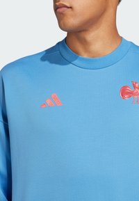 Mies, joka on pukeutunut vaaleansiniseen paitaan, jossa on punainen Adidas-logo ja punainen kukko embleemi rinnassa, kaula ja alaleuka näkyvät.