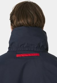 Marineblauwe jas met een hoge kraag, voorzien van een rode accentlijn op de achterkant. Glad, lichtgewicht materiaal met een getailleerde pasvorm.