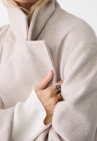 Persona che indossa un cappotto di lana beige con colletto alto e maniche di maglione bianco a costine, mostrando la mano con un tatuaggio di serpente e anelli d'oro.