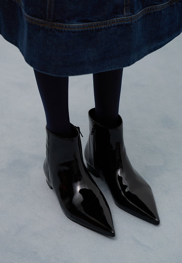 Czarne lakierowane botki za kostkę z ostro zakończonym noskiem, bocznym zamkiem błyskawicznym i elegancką sylwetką, zestawione z ciemnymi rajstopami i dżinsową spódnicą.
