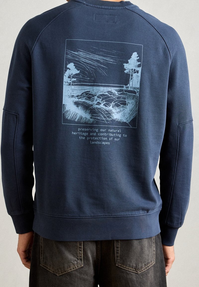 Felpa blu navy con maniche a raglan, caratterizzata da un'illustrazione paesaggistica stampata in azzurro e testo sul retro. Tessuto morbido in misto cotone.