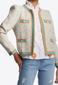 Veste en tweed crème avec des accents orange et bleu. Elle possède un col rond, deux poches et des boutons décoratifs. Portée par-dessus une chemise blanche brodée.