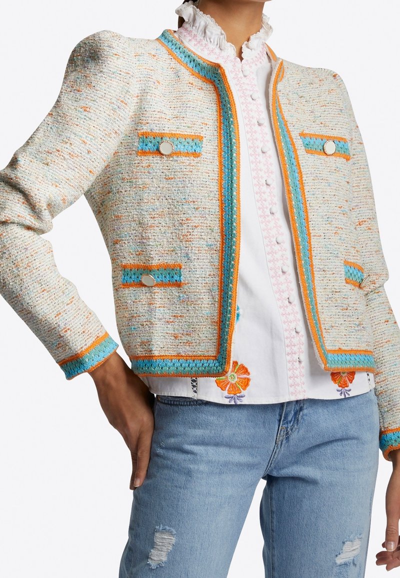 Veste en tweed crème avec des accents orange et bleu. Elle possède un col rond, deux poches et des boutons décoratifs. Portée par-dessus une chemise blanche brodée.