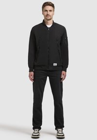 Schwarze Bomberjacke mit Reißverschlusstaschen, gerippten Bündchen und Saum, kombiniert mit dunklen Jeans und schwarz-weißen Turnschuhen.