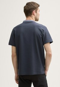 Polo a maniche corte di colore blu scuro con tessuto texturizzato e colletto in un blu chiaro a contrasto. La vista posteriore mostra un design semplice e una vestibilità comoda.