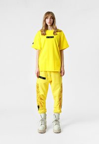 Camiseta de algodón amarilla de corte oversize con una etiqueta negra, combinada con pantalones amarillos a juego que presentan estampados artísticos y bolsillos laterales. Botas blancas.