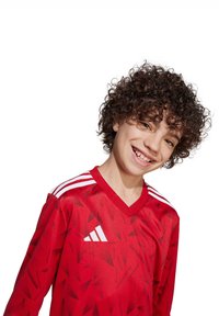Maglietta da calcio rossa con un motivo geometrico, presenta righe bianche Adidas sulle spalle e un design a V. Tessuto di texture liscia.