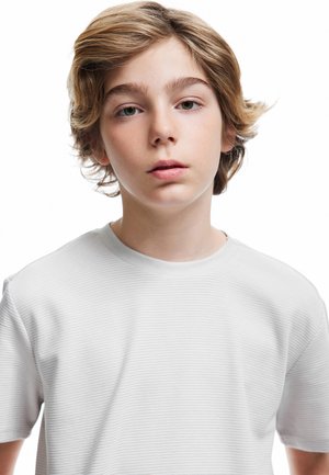 Hvid t-shirt med korte ærmer og en subtil ribbet tekstur, der har en rund halsudskæring. Modellen har mellemlangt bølget hår og et neutralt udtryk.