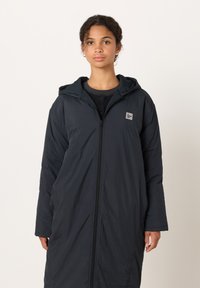 Ung kvinna med lockigt hår, iklädd en lång svart huva med dragkedja över en mörk tröja, stående framför en slät ljus bakgrund.