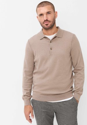 Man met kort haar en baard, die een beige lange mouwen polotrui draagt over een wit overhemd en grijze geruite broek tegen een witte achtergrond.