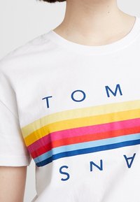 Biała bawełniana koszulka z wyraźnymi poziomymi paskami w kolorach żółtym, różowym, czerwonym i niebieskim, z nadrukiem "TOM" i "SNA" w granatowym kolorze.