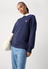 Ναυτικό φούτερ με στρογγυλή λαιμόκοψη, ριμπ μανίκια και στρίφωμα, και ένα μικρό κόκκινο λογότυπο στο στήθος. Συνδυάζεται με ανοιχτό μπλε τζιν και άσπρη τσάντα.