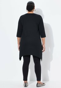 Zwarte tuniek met driekwart mouwen, hoge-lage zoom, gecombineerd met zwarte leggings en gegolfde platte schoenen. Glad materiaal met een lichte glans.