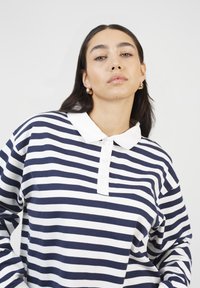 Woman with long dark hair wearing gold hoop earrings and a navy and white striped long-sleeve shirt with a white collar on a white background.