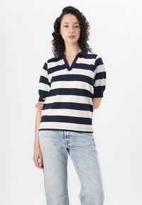 Marineblauw en crème gestreepte polo met een v-hals en korte pofmouwen; gemaakt van een zachte stof. Gecombineerd met lichtblauwe spijkerbroeken.