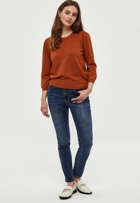 Pulover u boji hrđe s rukavima do tri četvrtine, uparen s plavim skinny trapericama i bijelim mokasinkama s zlatnim detaljima. Glatka tekstura.