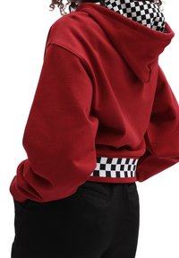 Persona vestida con una sudadera roja con cinturilla y interior de capucha a cuadros en blanco y negro, combinada con pantalones negros, con las manos en los bolsillos, de espaldas.