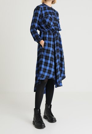 Kobieta ubrana w długą sukienkę w niebiesko-czarną kratę z wciętą talią, czarne legginsy i czarne sznurowane buty, stojąca z rękami w kieszeniach.