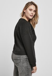 Felpa nera con pannelli in pizzo testurizzato sulle spalle, colletto rotondo e orlo a coste. Abbinata a jeans grigi aderenti, che evidenziano un look casual.