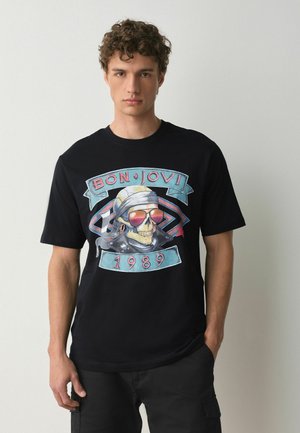 Ung mand iført en sort Bon Jovi 1989 t-shirt med en kranie med solbriller og bandana, stående mod en ensfarvet lys baggrund.
