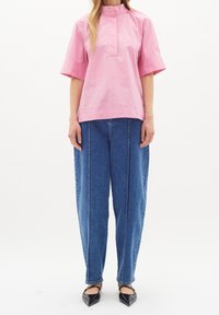 Kvinna i en lös rosa kortärmad skjorta, avslappnade blå jeans med framtilliggande sömmar och svarta spetsiga flats med remmar, stående.